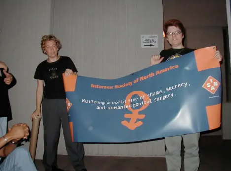 Thea and Bo, two white intersex people, stand holding a banner that says ISNA: Building a world free of shame, secrecy, and unwanted genital surgery 