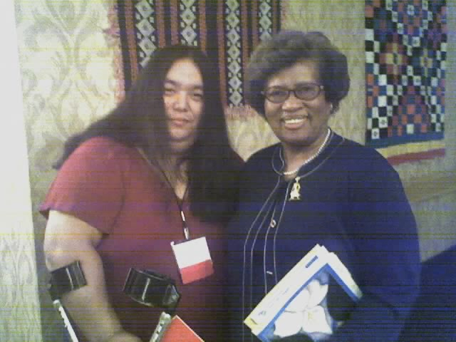 Emi, wearing a conference lanyard, stands using an arm crutch, smiling, next to Jocelyn Elders