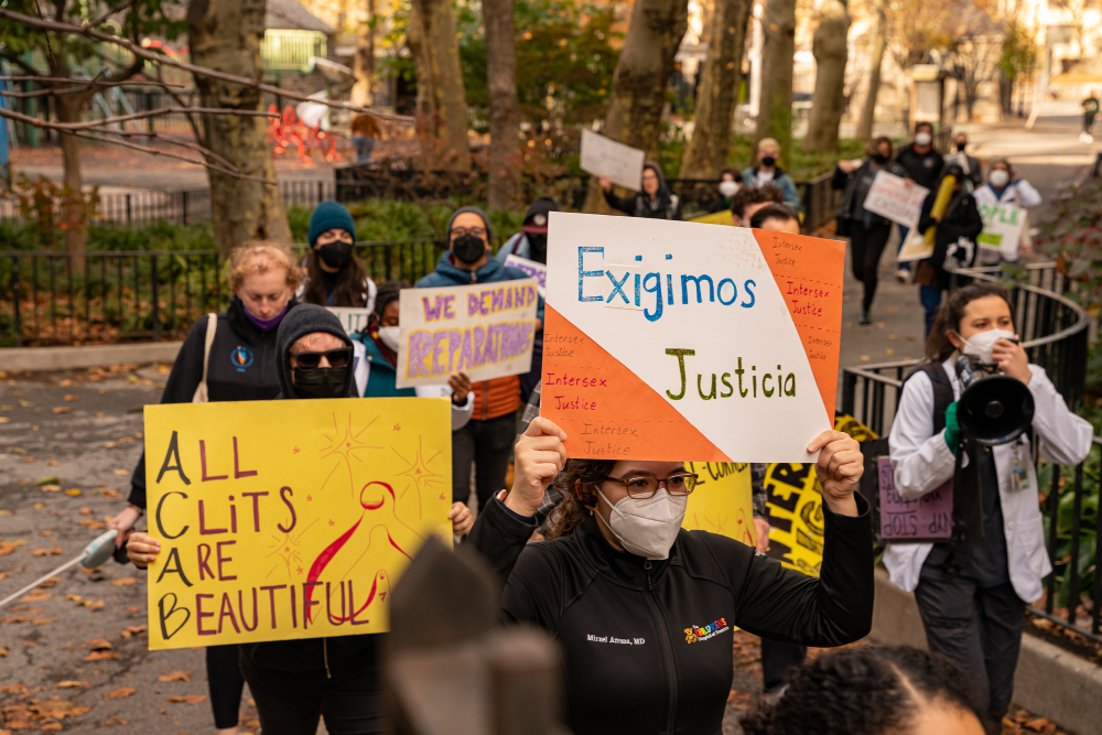 People in N95 masks march through a winding park path covered in autumn foliage, holding signs including All Clits Are Beautiful