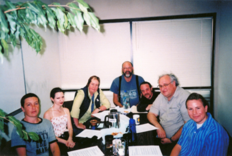 Six people sit around a library table, stopping for a photo with coffee cups and papers spread out