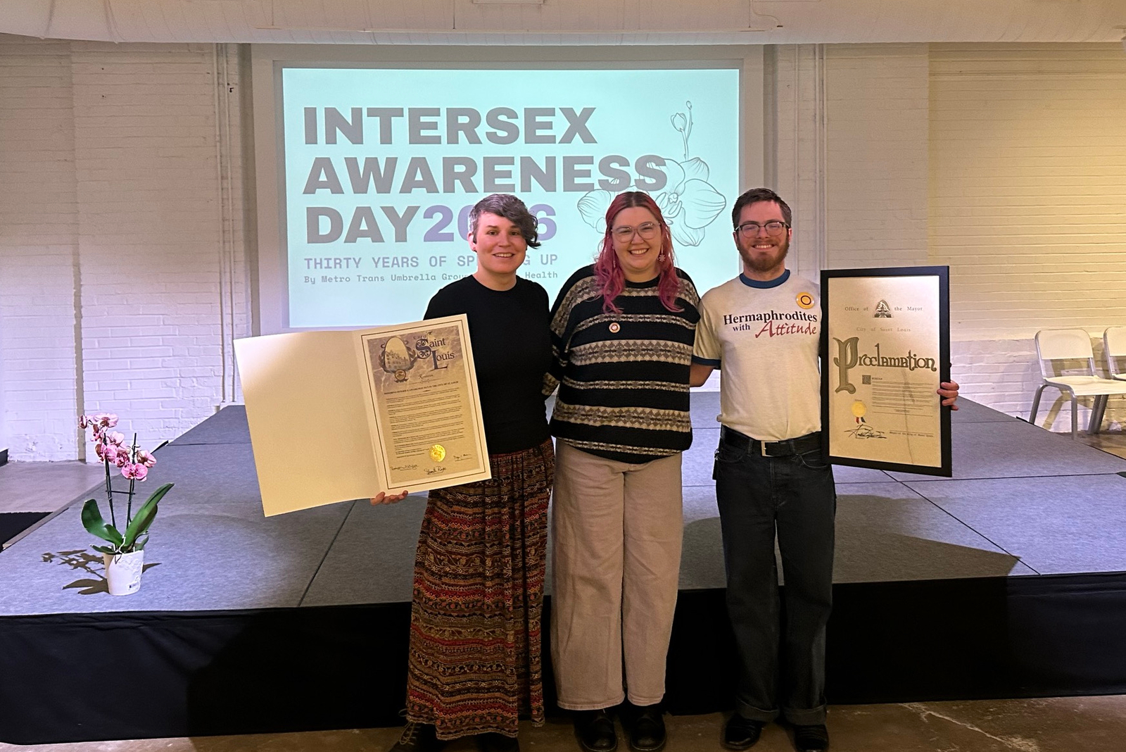 Advocates stand inside a conference room in front of a projector screen holding up copies of the city's resolution and proclamation in frames
