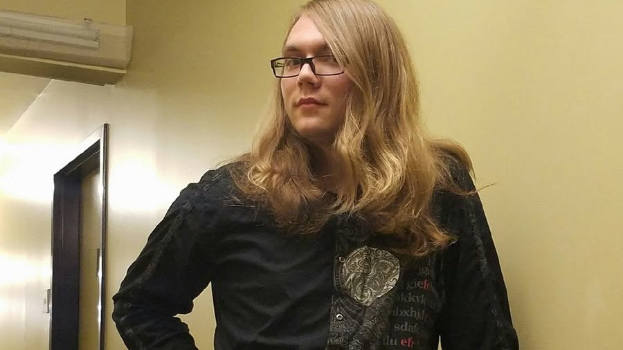 Scout Schultz standing in a dorm hallway, shoulder-length blonde hair and glasses, wearing a black dress shirt