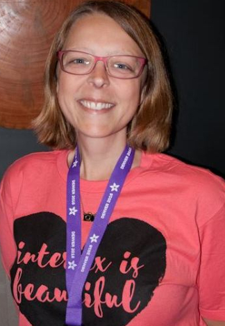 Amy, a white intersex woman with a blonde shoulder length bob and glasses, smiles wearing a purple intersex conference lanyard and one of Pidgeon's shirts, "intersex is beautiful" on a heart design