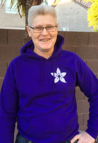 Jim, a white intersex man with white hair and glasses, smiles facing forward with one hand on his hip. He wears a bright purple hoodie from the AISDSD intersex conference.