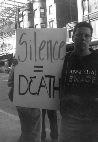 Black and white photo of Max at a protest holding up a Silence = Death sign. Max wears a Transsexual Menace t-shirt.